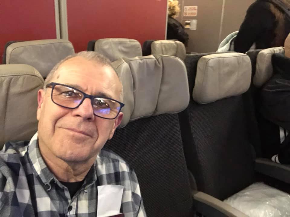 Mick, a grandfather aged white guy is smiling at the camera from a coveted aisle seat of the plane. He’s wearing a black and white checked shirt with a black T-shirt underneath and black glasses frames. He’s so coordinated he even blends in with the black seats!