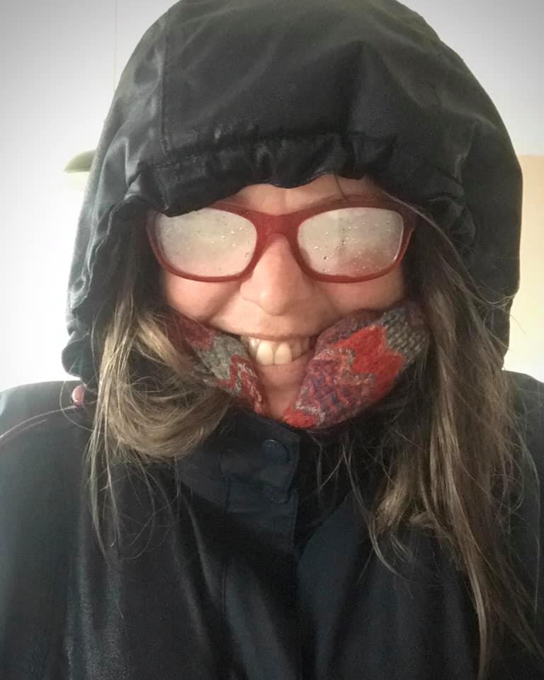 Caitlin, a white woman, has been caught in a storm - her red glasses are all steamed up and her blue waterproof coat and hood look black because it’s so wet!