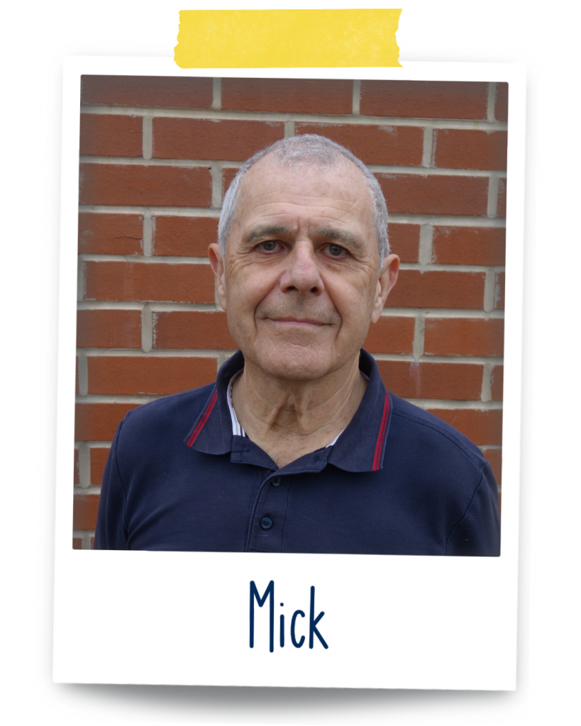 Mick, a 72 year old, UK white British man with short grey hair is  wearing a navy blue golf t-shirt. He's standing against a brick wall slightly smiling looking at the camera.