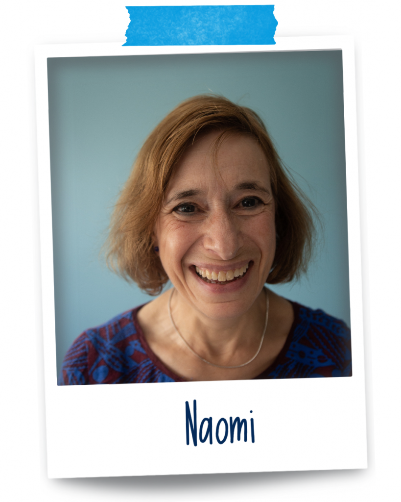 Naomi is a white woman in her 60s with wavy mid-brown hair and grey eyes. She is wearing a brown and blue top, a necklace, and has a bright smile on her face.