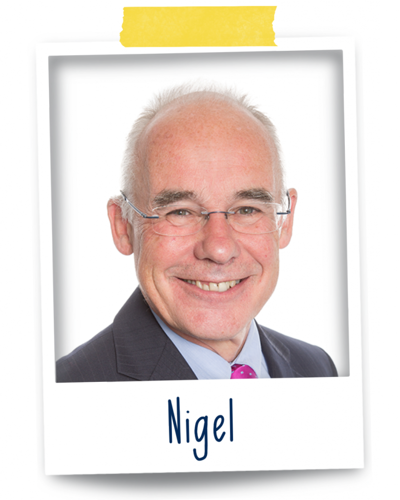 Nigel,  a 70 year old glasses-wearing man with, at best, limited short grey hair is smiling at the camera wearing a faintly stripped black suit, light blue shirt and a pink tie.